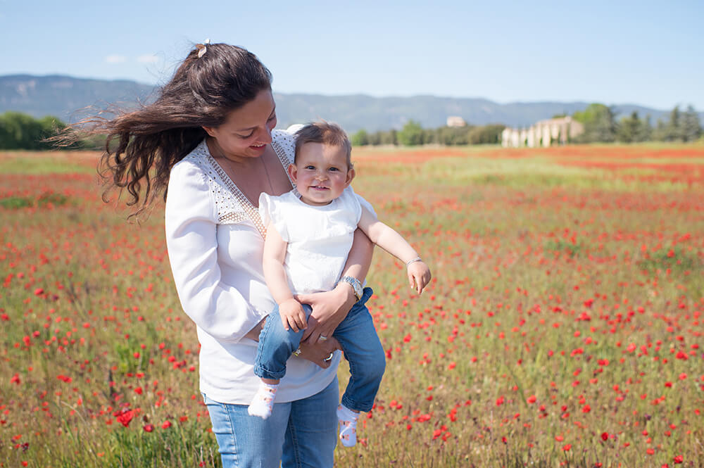 Portrait de bébé d'un an dans les bras de sa maman et au milieu d'un énorme champ de coquelicots.