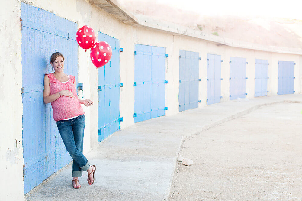 Femme enceinte tenant des ballons rouges hélium devant des portes bleues.