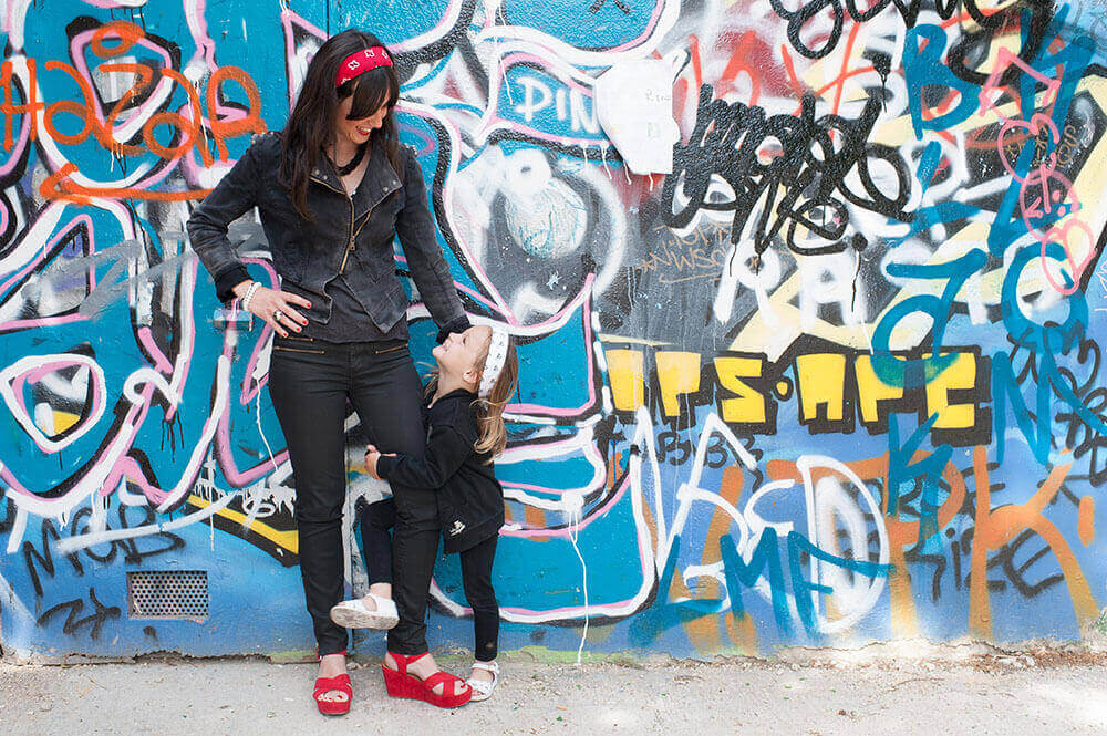 Devant un mur de graffitis portrait d'une enfant qui tient la jambe de sa maman
