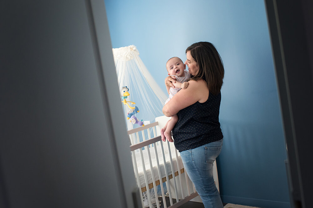 Bébé dans les bras de sa maman à côté de son lit.